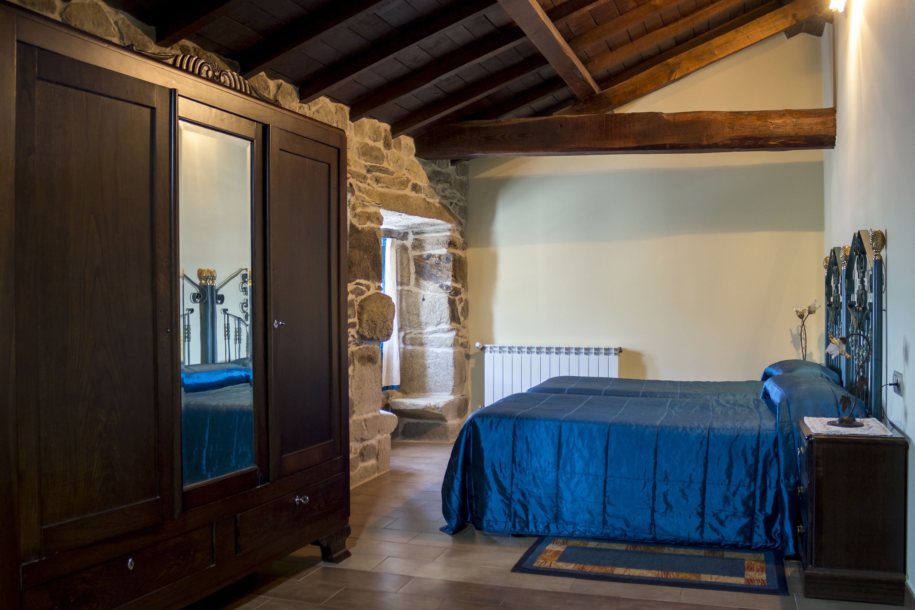 Une chambre avec un mur en pierre, des poutres apparentes en bois au plafond, une grande armoire et un lit recouvert d’une couverture bleue.