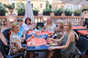 Un grupo de personas cenando en una mesa al aire libre, con plantas en macetas y suelo de baldosas.