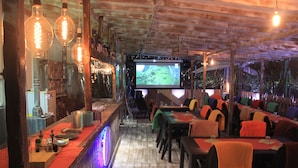 A restaurant with a bar area, tables with orange and green tablecloths, and hanging light bulbs. - The Roma House (Rome)