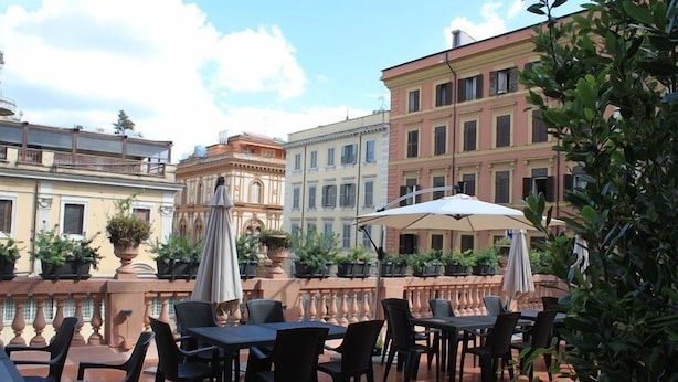 An outdoor patio with tables and chairs, surrounded by buildings with balconies and potted plants. - The Roma House (Rome)