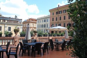 An outdoor patio with tables and chairs, surrounded by buildings with balconies and potted plants. - The Roma House (Rome)