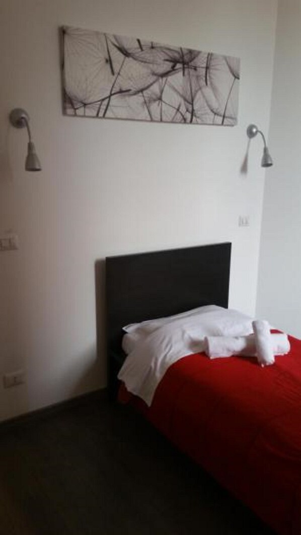 A bedroom with a black headboard, a red bedspread, and a white towel on the bed. There is a wall-mounted abstract art piece above the bed.