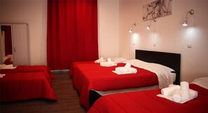 Habitación de hotel con dos camas, cada una con ropa de cama roja y toallas blancas. Se observan luces fijadas en la pared y una puerta.