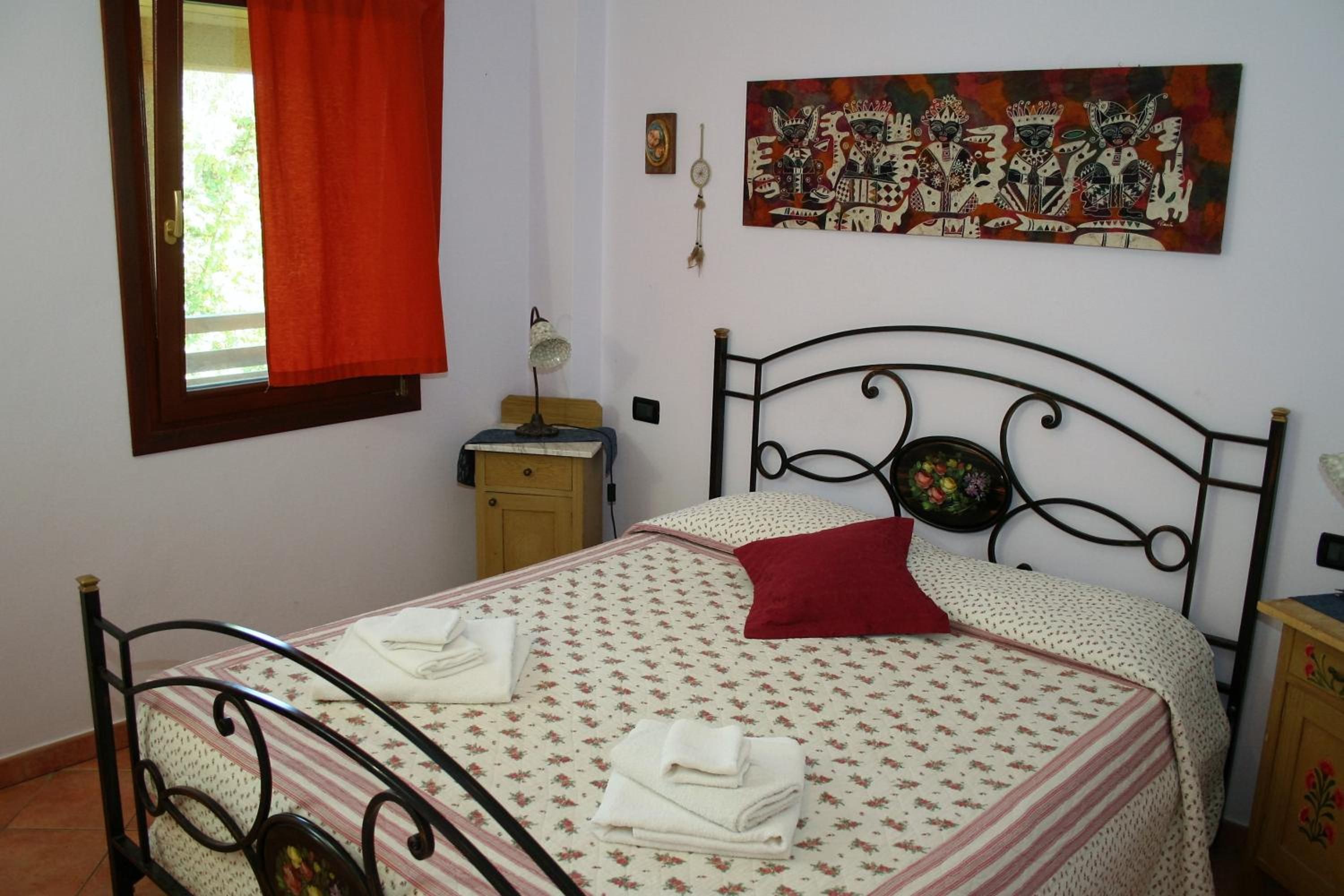 Un dormitorio con cama, mesita de noche, lámpara, un cuadro y una ventana con cortinas.