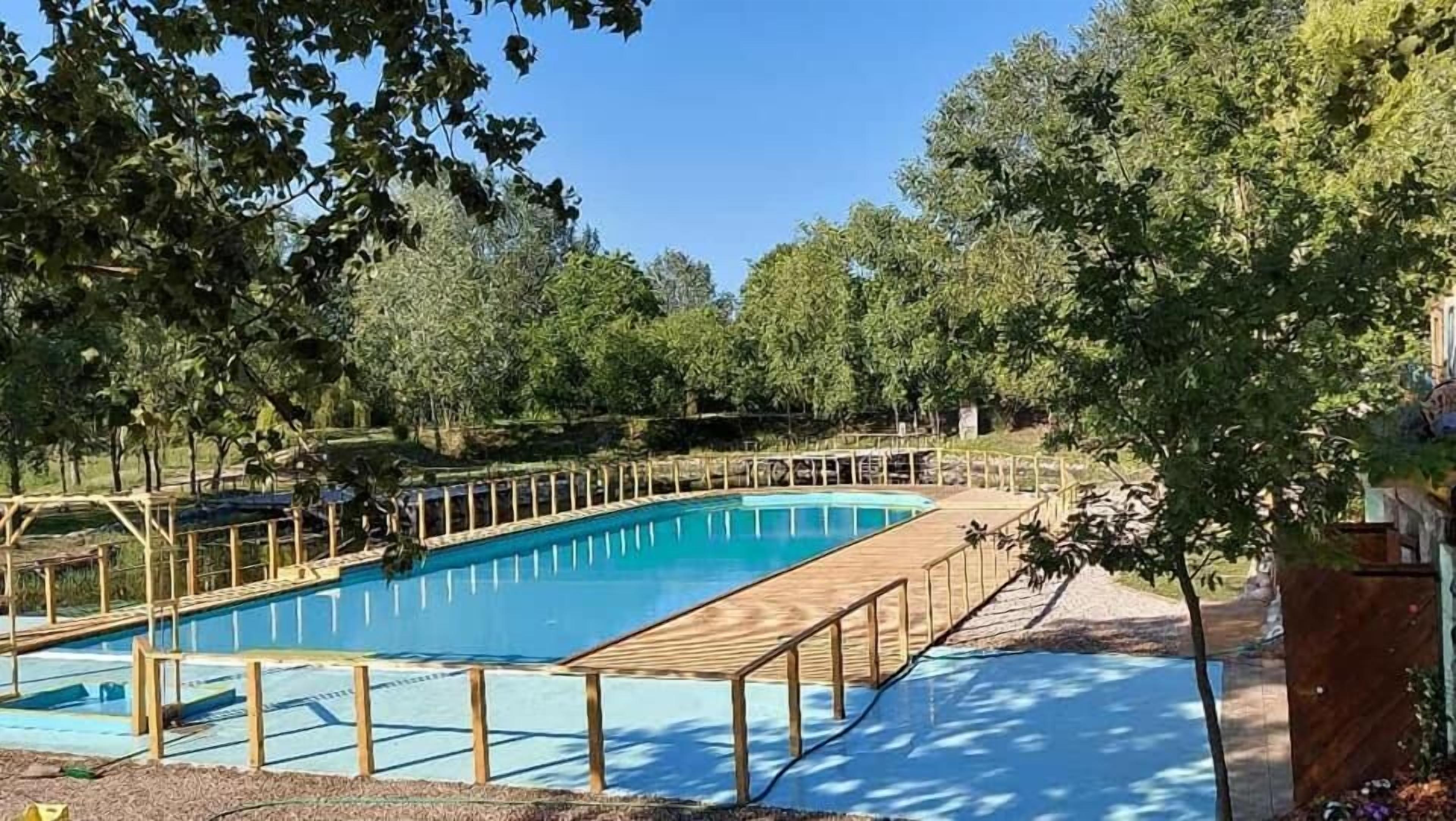 Una piscina rettangolare con bordo in legno e ringhiere di sicurezza, circondata da alberi.