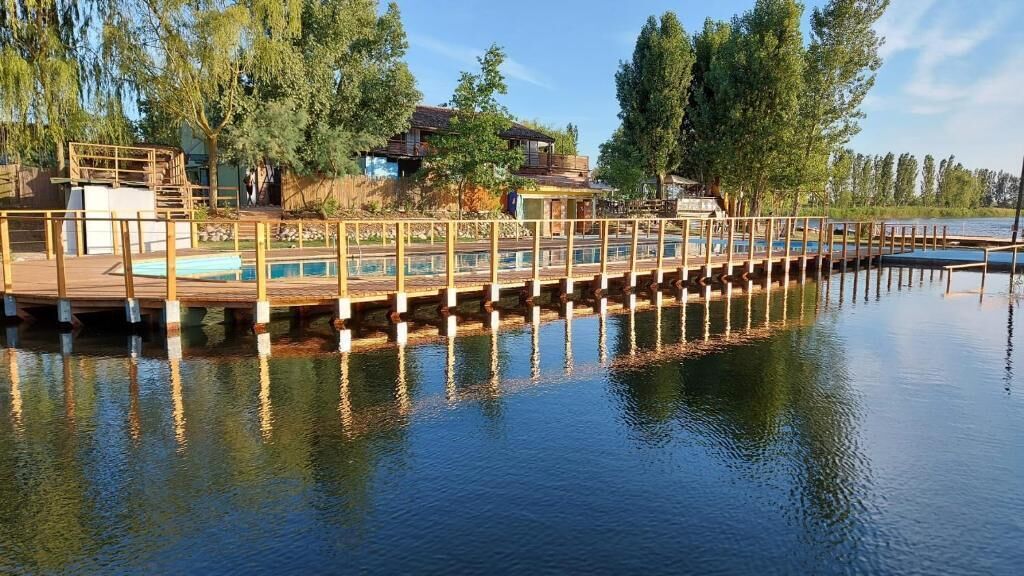 Eine hölzerne Anlegestelle erstreckt sich über ruhiges Wasser, umgeben von Bäumen und einem Gebäude mit einer Terrasse.