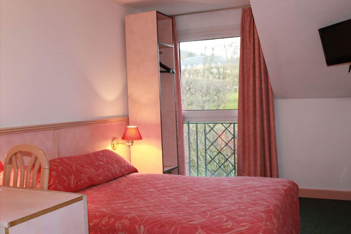 Ein Hotelzimmer mit einem Bett, einem Sessel, einem Schreibtisch, einem Fenster mit Vorhängen und einem an der Wand befestigten Fernseher.