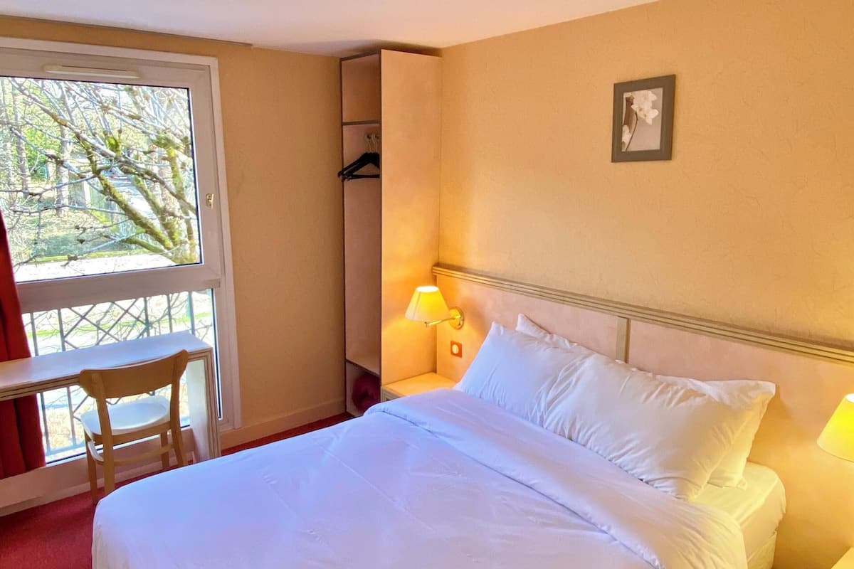 Ein Hotelzimmer mit Bett, Sessel, Nachttisch, Lampe und Fenster mit Blick auf Bäume.