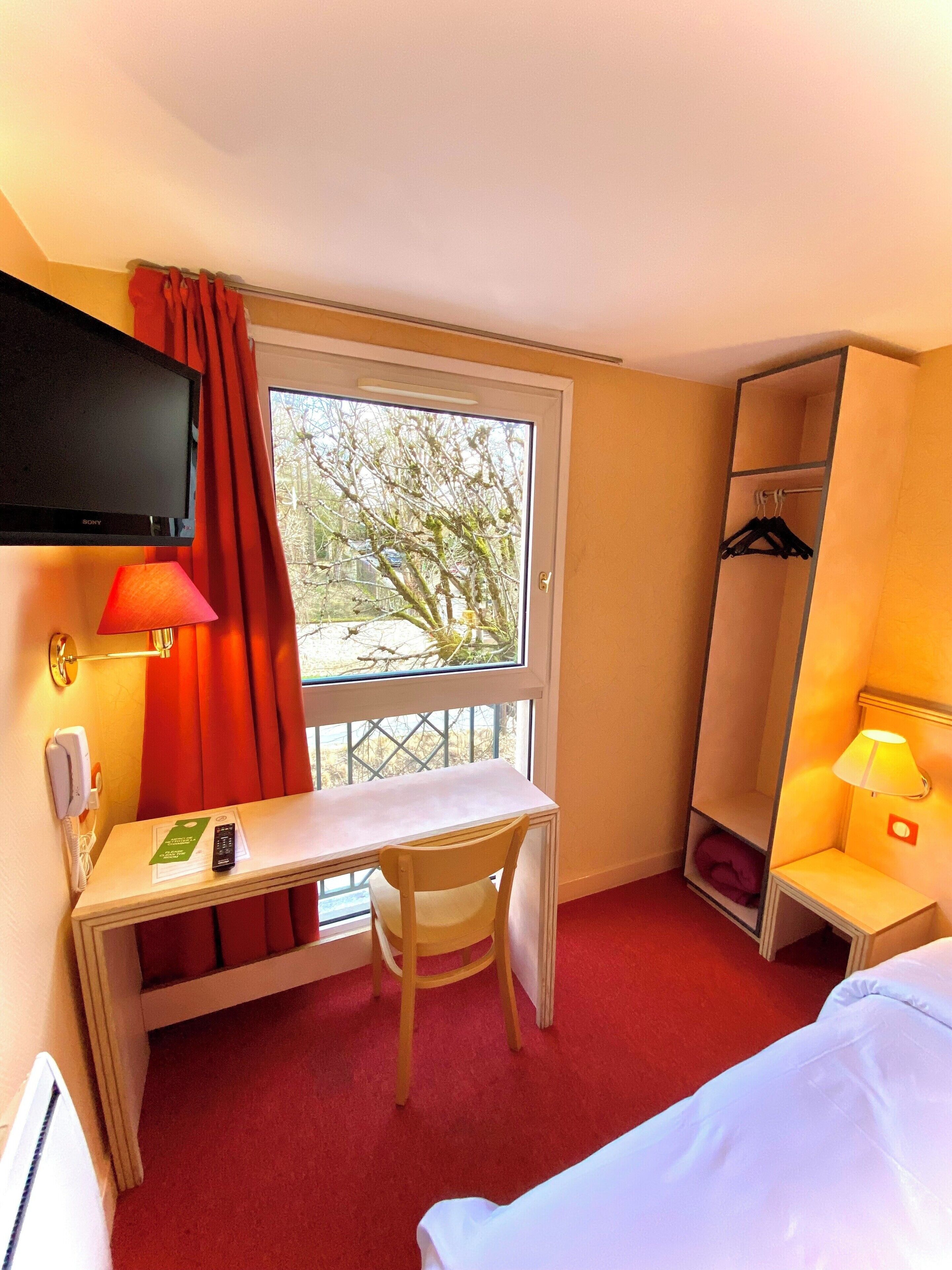 Une chambre d’hôtel équipée d’un bureau, d’une chaise, d’un lit, d’une télévision et d’une fenêtre donnant sur des arbres.