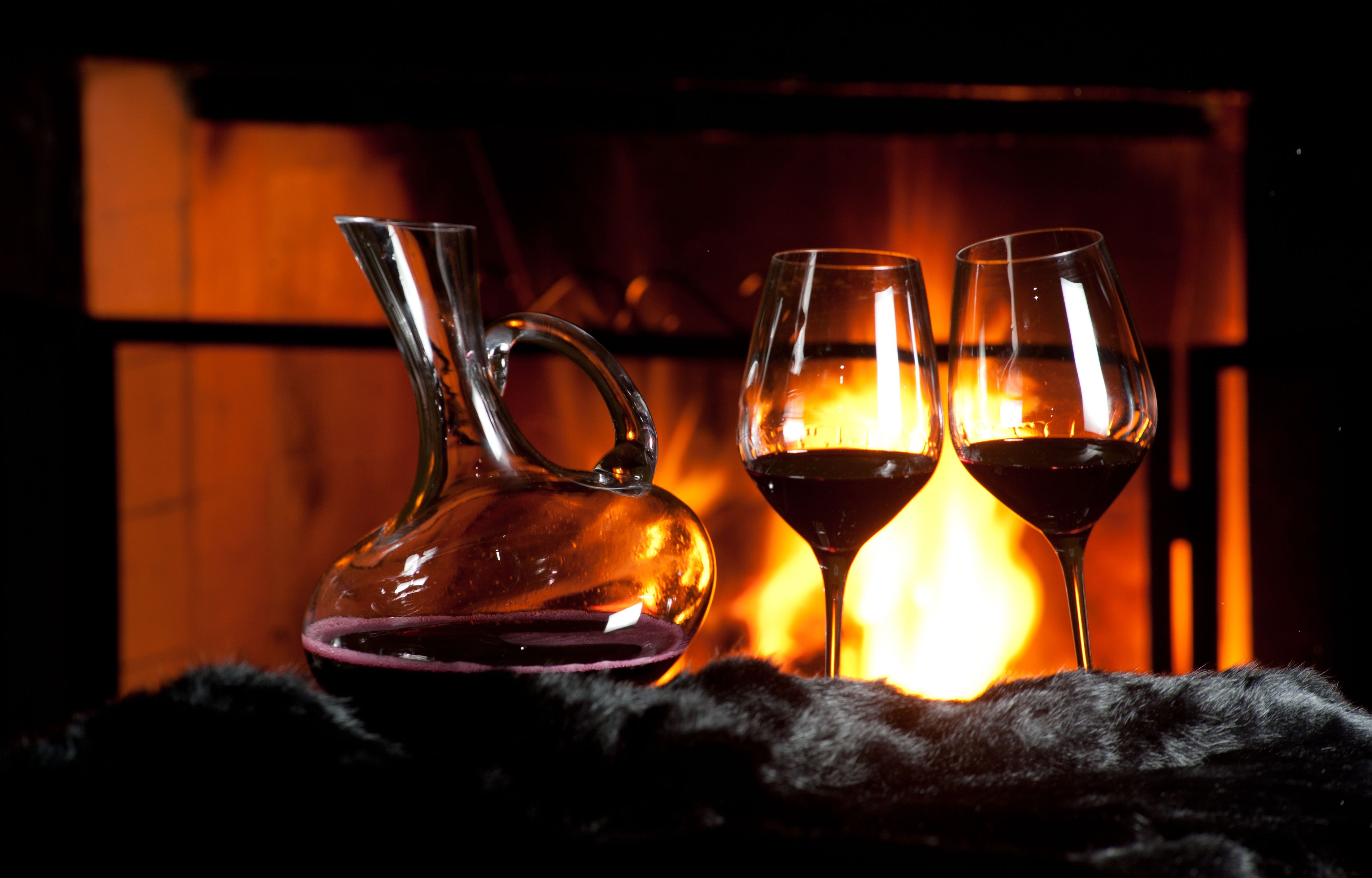 A glass decanter and two wine glasses on a dark surface with a fireplace in the background.