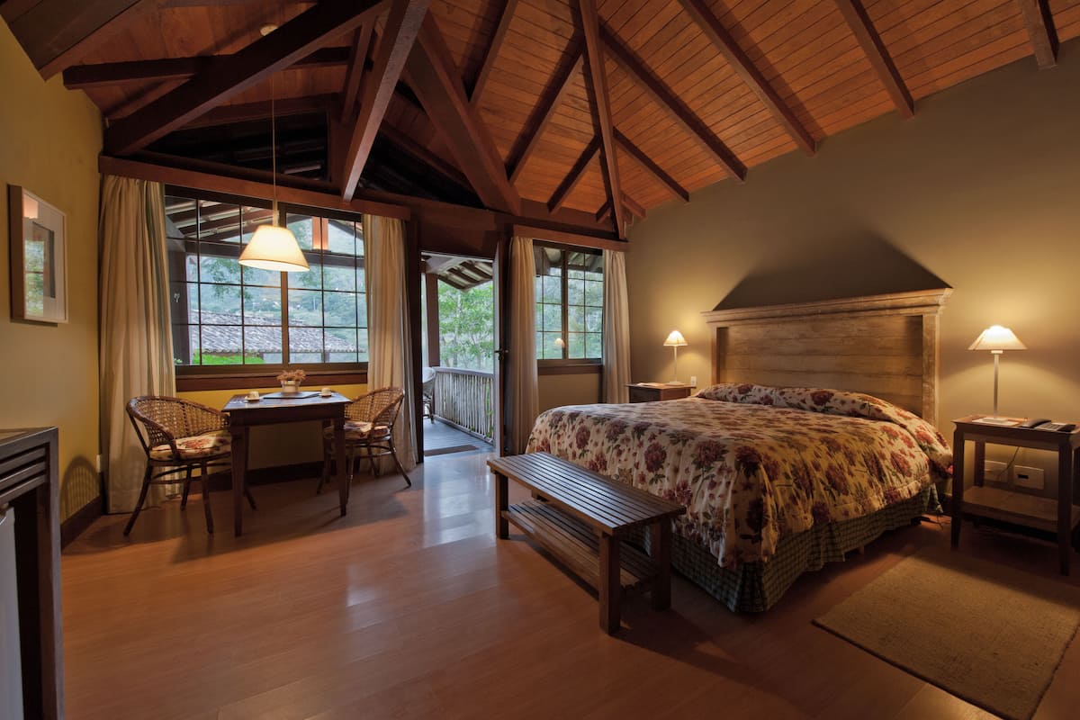 Ein gemütliches Schlafzimmer mit Holzbalkendecke, einem großen Bett mit floralem Bettzeug, einem Nachttisch mit Lampe und Blick ins Freie durch ein Fenster.