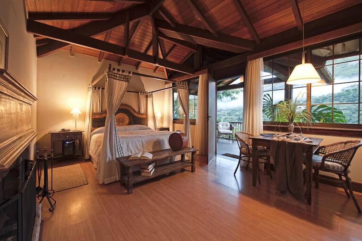 Ein geräumiges Schlafzimmer mit einem Himmelbett, einem Holztisch und Stühlen, einem Kamin und großen Fenstern mit Blick ins Freie.