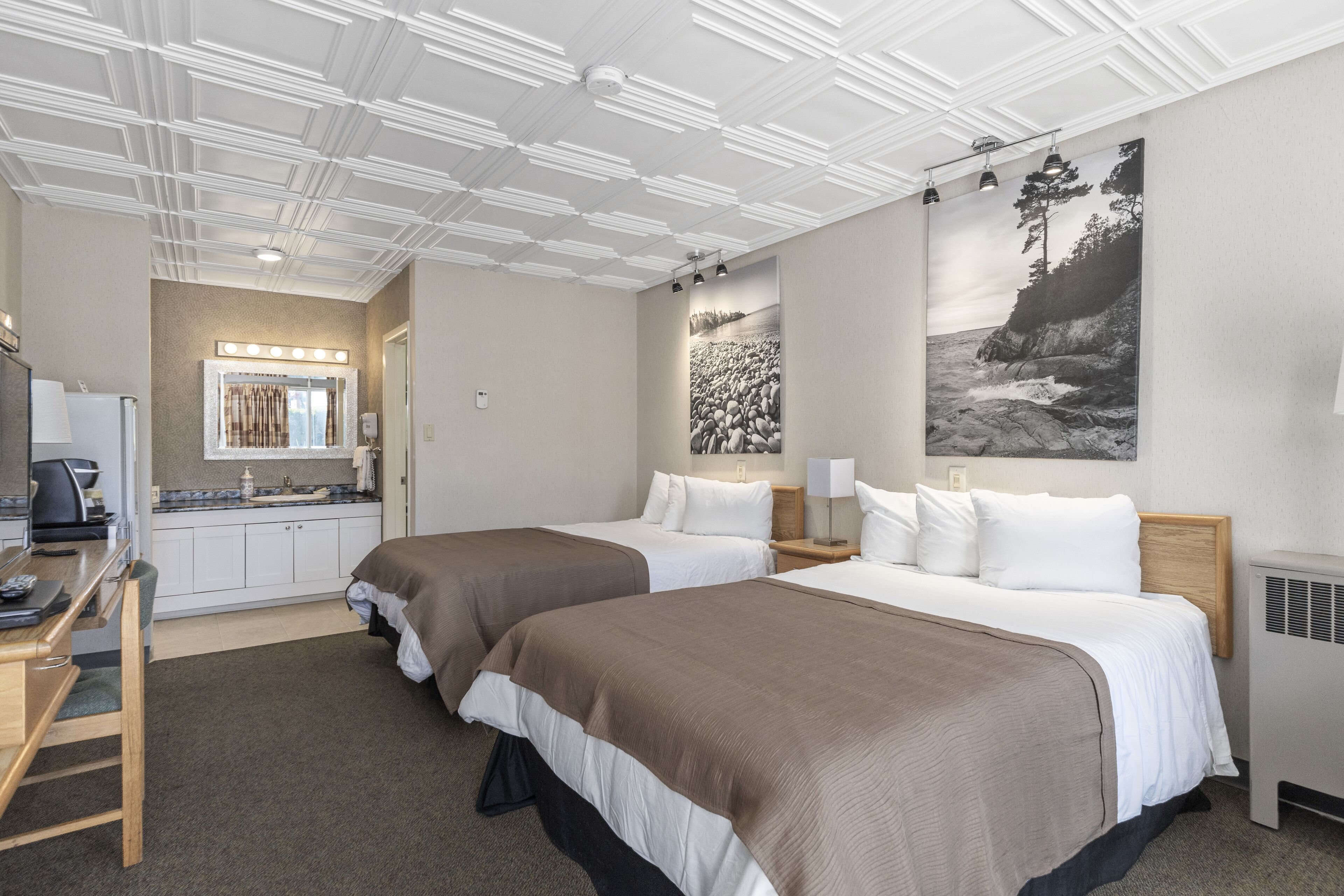 Une chambre d’hôtel avec deux lits, une kitchenette, une salle de bain avec un miroir et des luminaires, et des œuvres d’art accrochées aux murs.