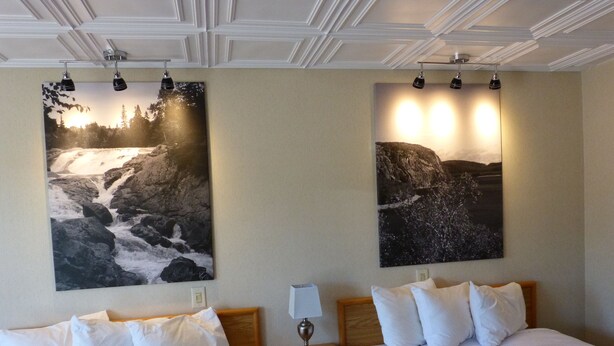 A hotel room with two beds, a nightstand, a lamp, and two large landscape paintings on the walls. - Catalina Motel (Sault Ste. Marie)