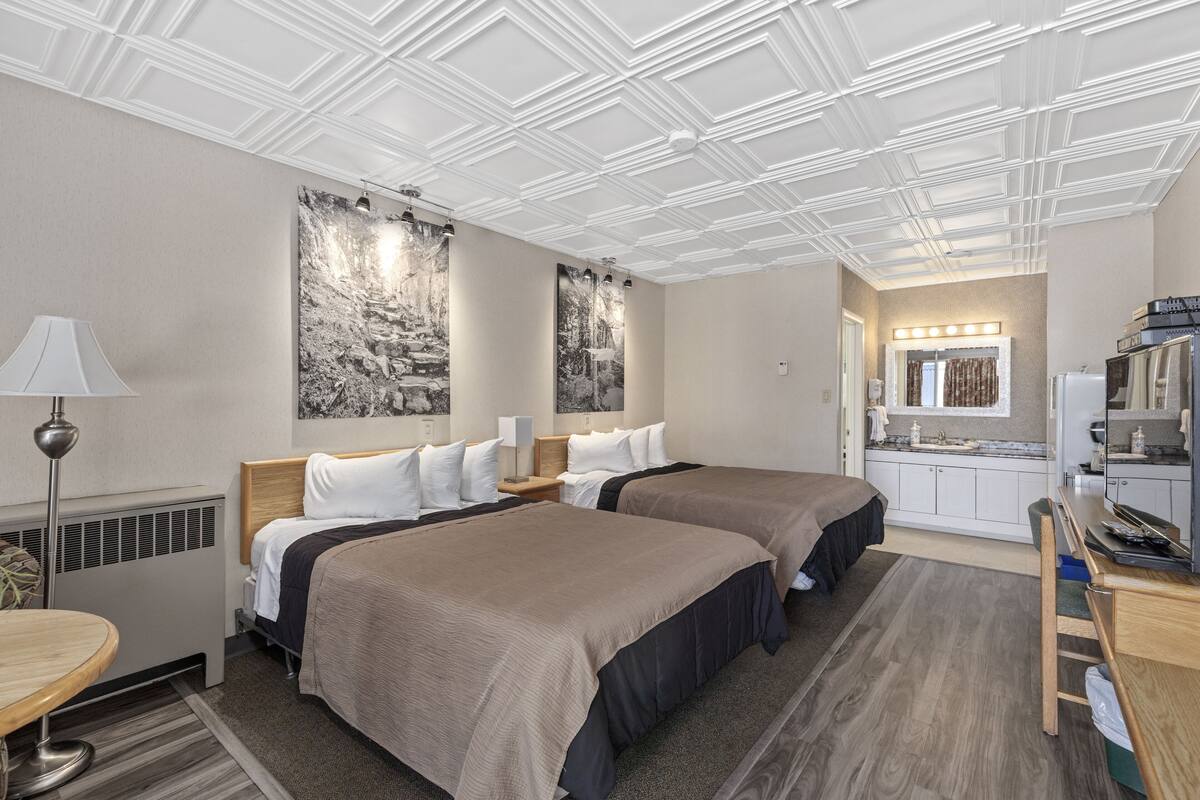 A hotel room with two beds, a desk, a chair, a lamp, a mirror, and a kitchenette.