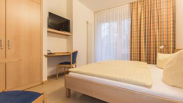 Une chambre à coucher avec un lit, un bureau avec une chaise, un téléviseur fixé au mur et une fenêtre avec des rideaux.
