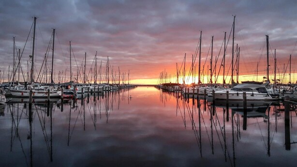 A marina with multiple boats docked, calm water reflecting the boats, and a sunset in the background. - Ferienpark Plauer See (Alt Schwerin)