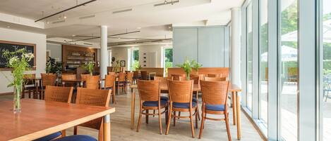 A spacious dining area with wooden tables and chairs, large windows offering views of the outdoors, and a bar area in the background.