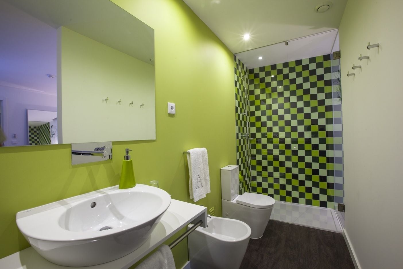 Un baño moderno con pared de ducha a cuadros verdes y blancos, un lavamanos blanco y un inodoro.