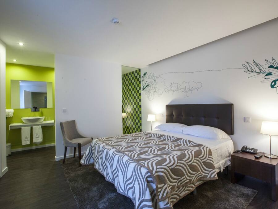 Habitación de hotel con cama, mesita de noche, silla y baño visible a través de una puerta abierta.