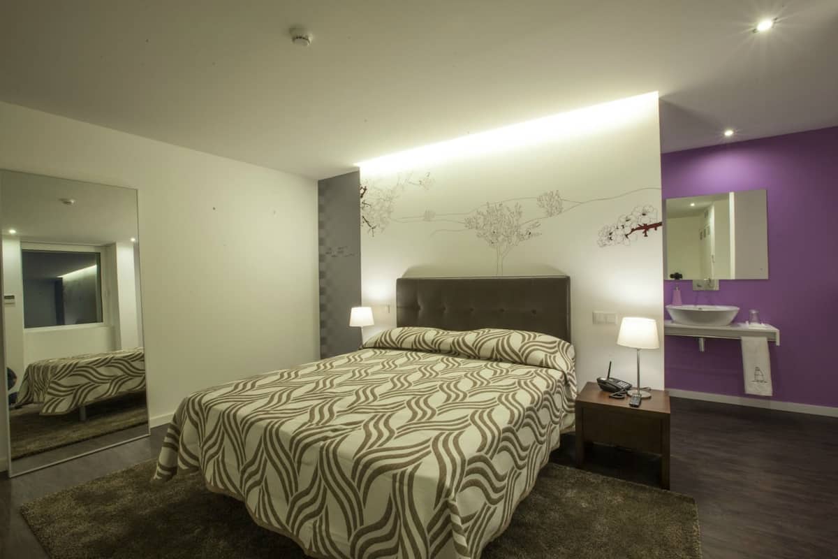 Ein modernes Schlafzimmer mit einem großen Bett, einem Nachttisch mit Lampe, einem Spiegel und einem Badezimmer mit Waschbecken und Spiegel.