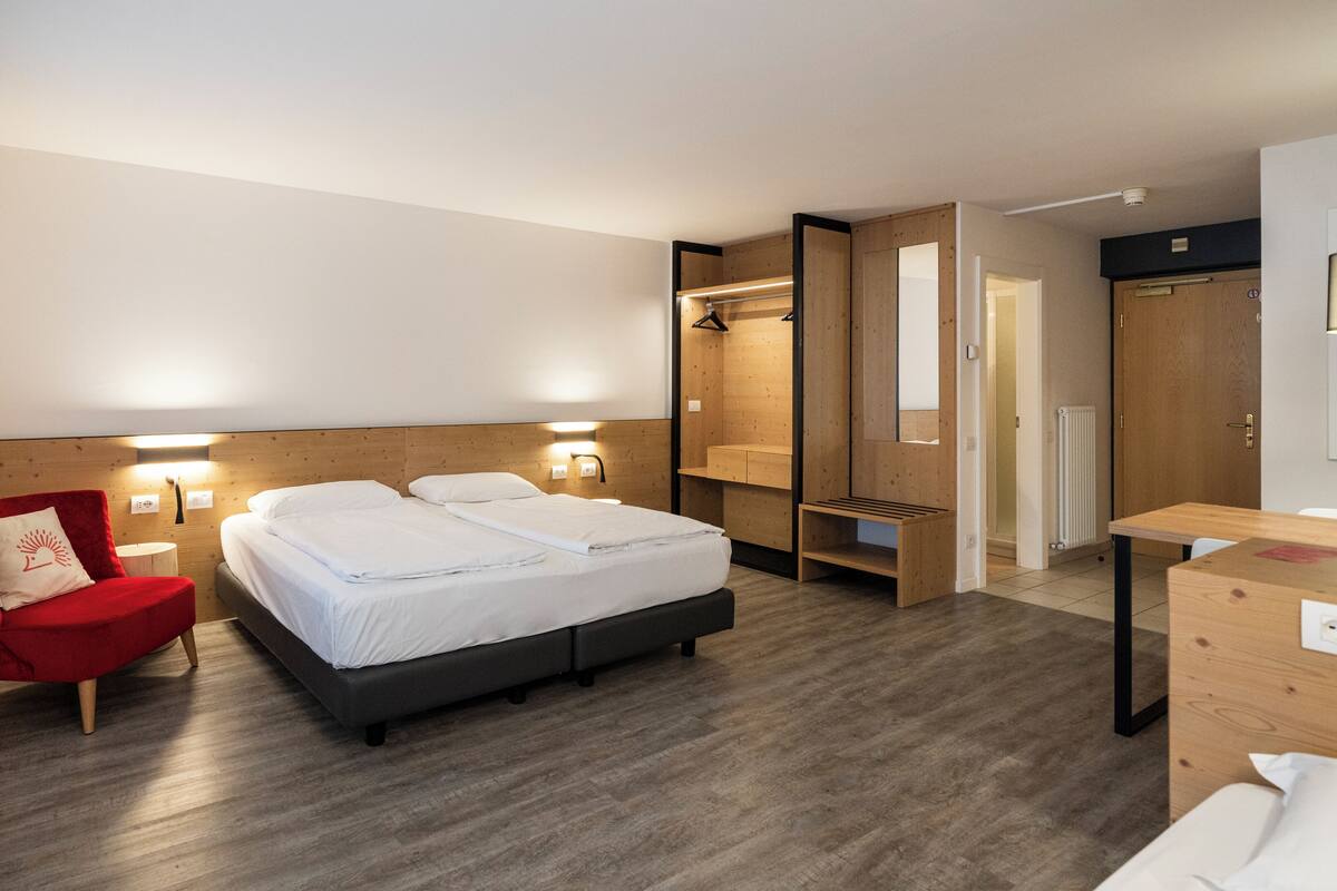 A modern hotel room with a double bed, a red chair, a wooden desk, and a mirror.