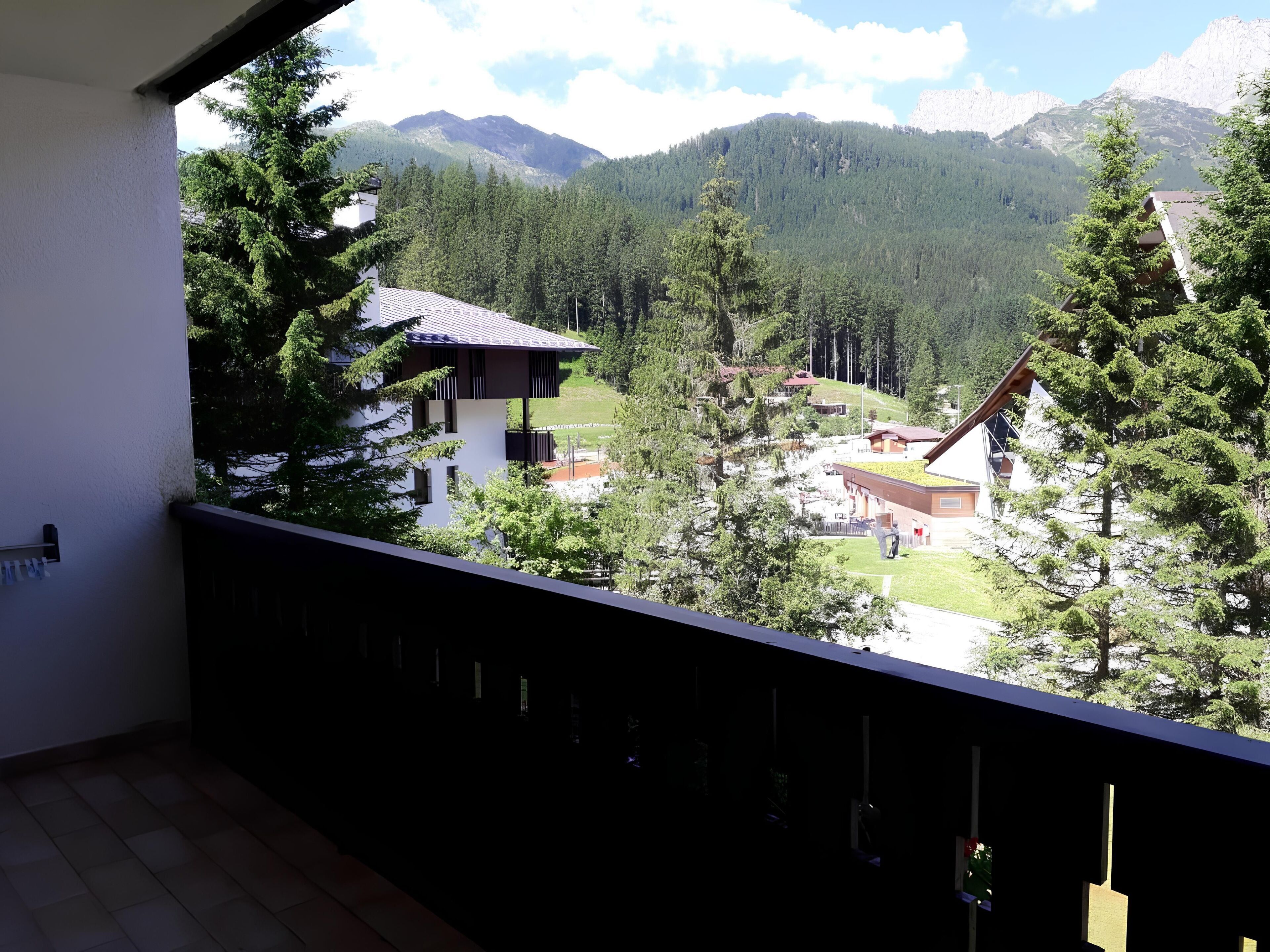 A balcony with a view of a mountainous landscape, a building with a sloped roof, and a person standing in the distance.