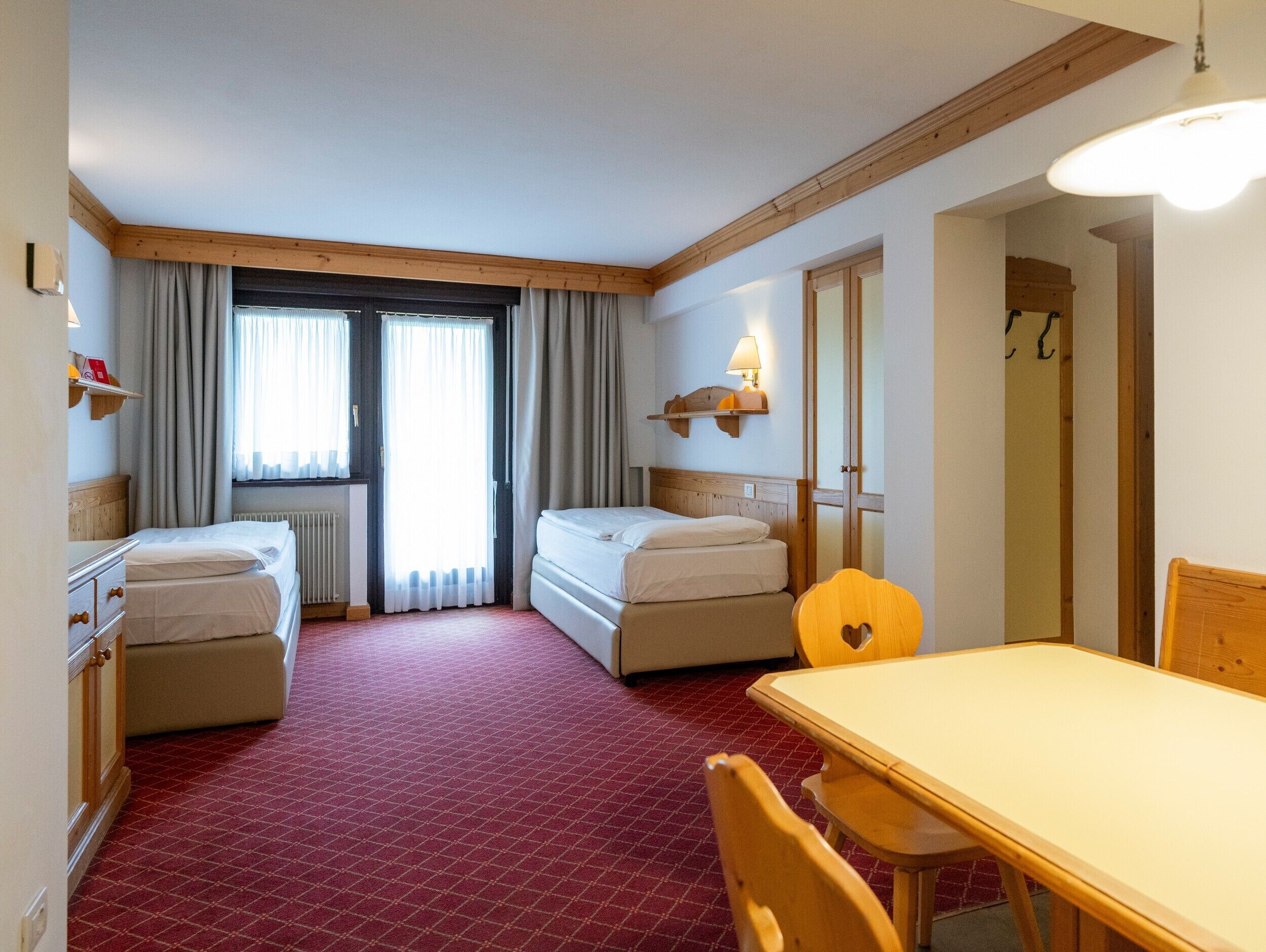 A hotel room with two beds, a dining table, a chair, a lamp, and a window with curtains.
