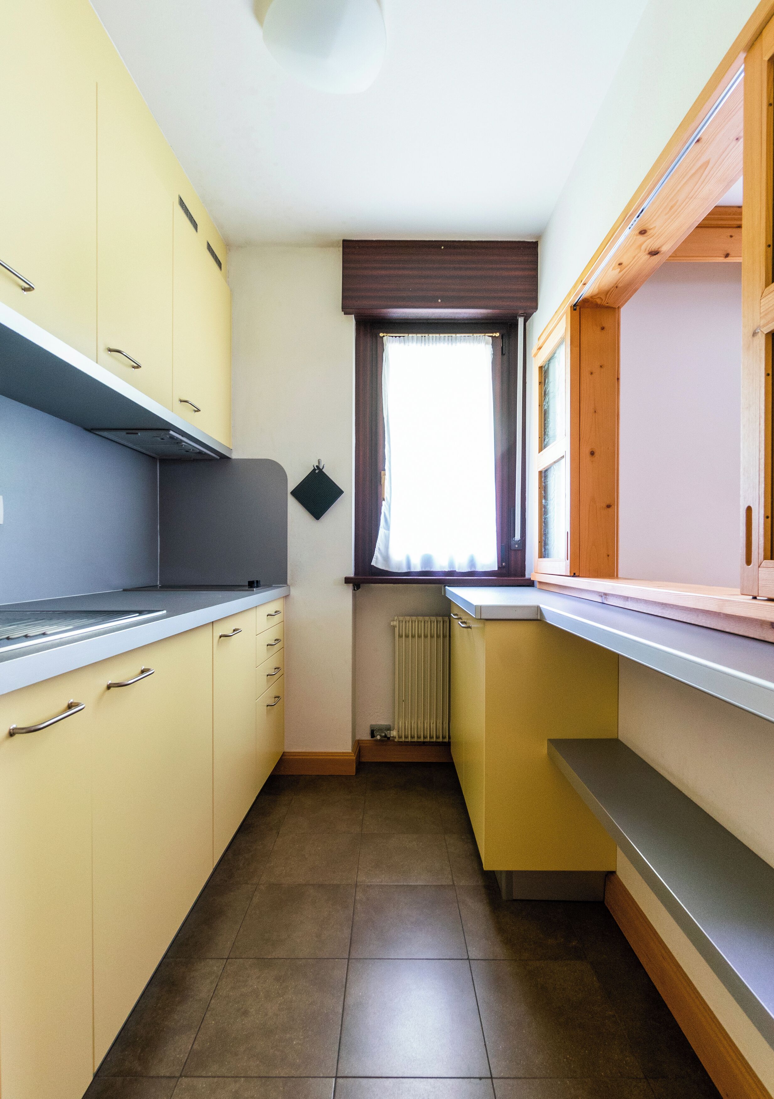 Eine schmale Küche mit gelben Schränken, einem Fenster mit Jalousien und einer Heizkörper.