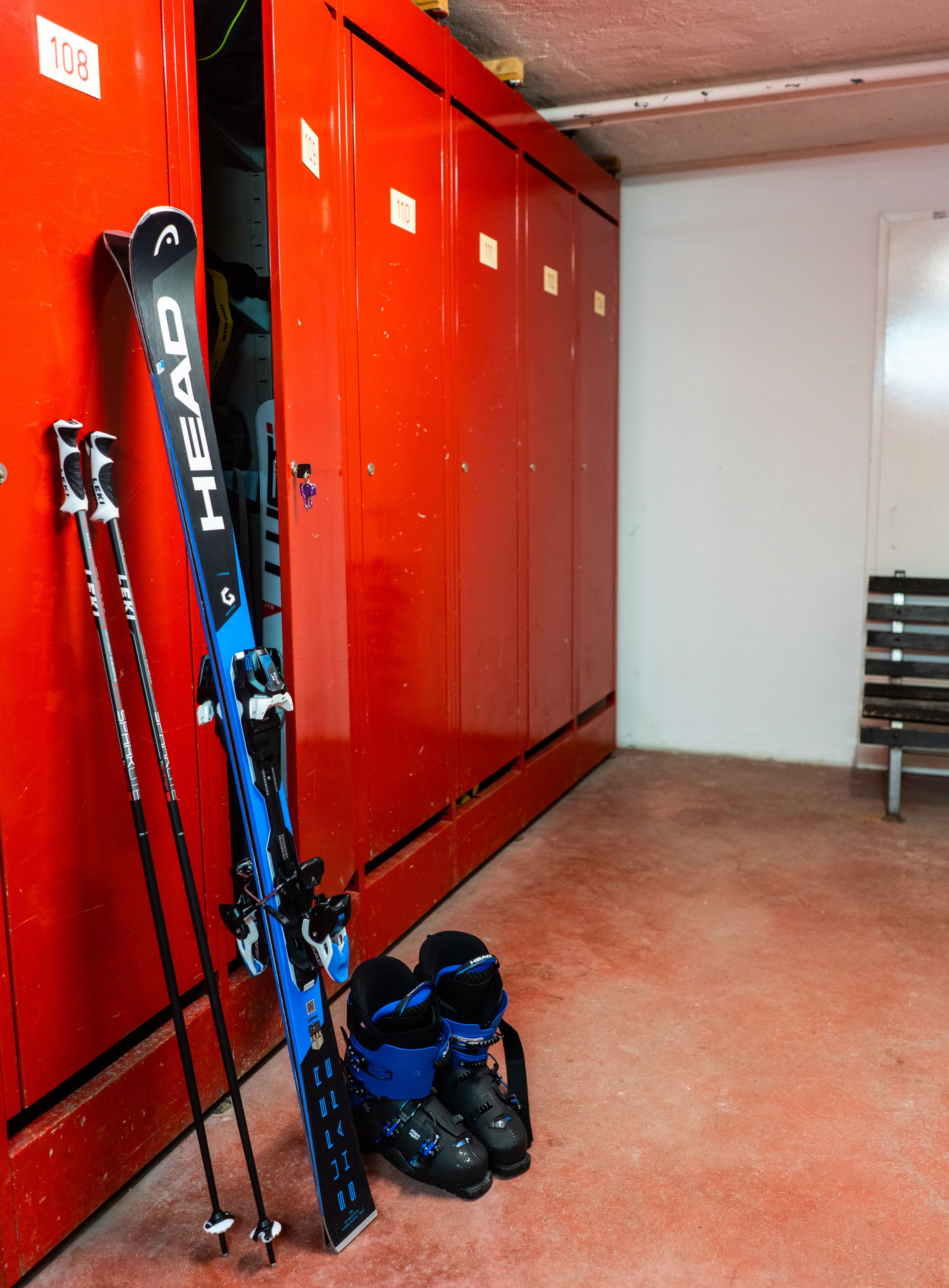 Ein roter Umkleideraum mit nummerierten Schließfächern, Ski, die an der Wand lehnen, und Skistiefeln auf dem Boden.