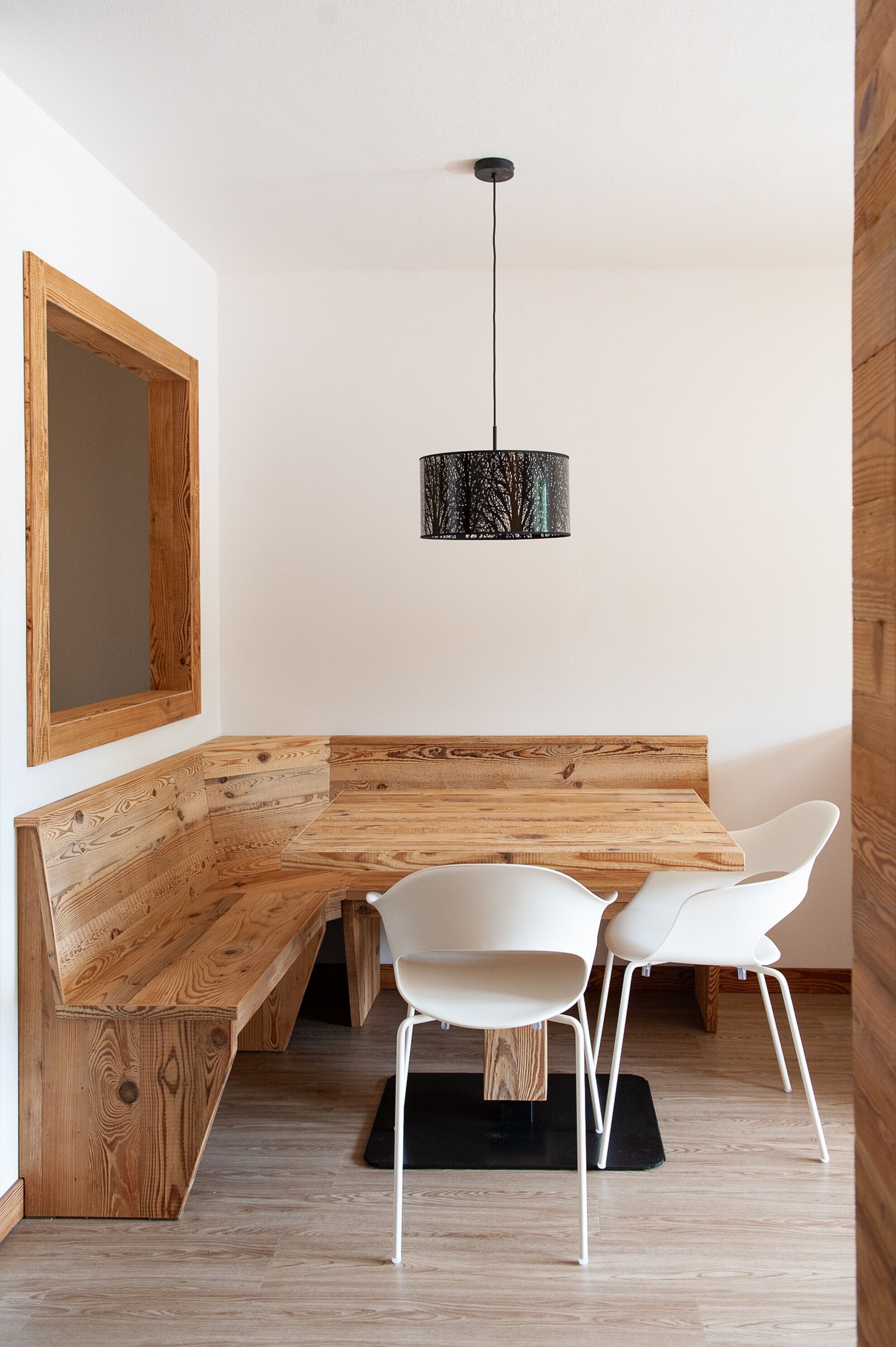 A dining area with a wooden bench and white chairs, a hanging pendant light, and a large mirror on the wall.