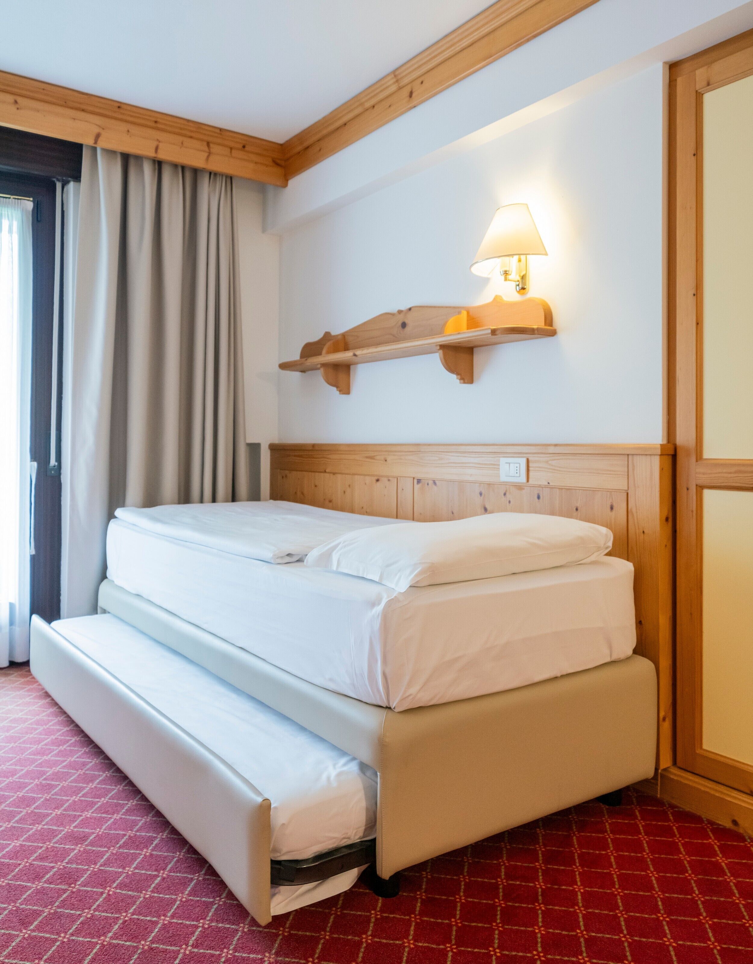 A hotel room with a bed, a wooden headboard, a wall-mounted lamp, and a red patterned carpet.