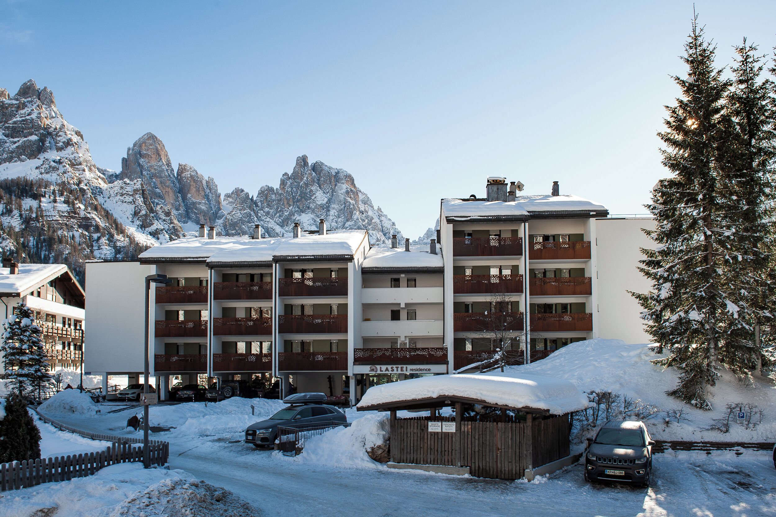 Un hotel de varios pisos con balcones, una zona de estacionamiento y una montaña nevada al fondo.