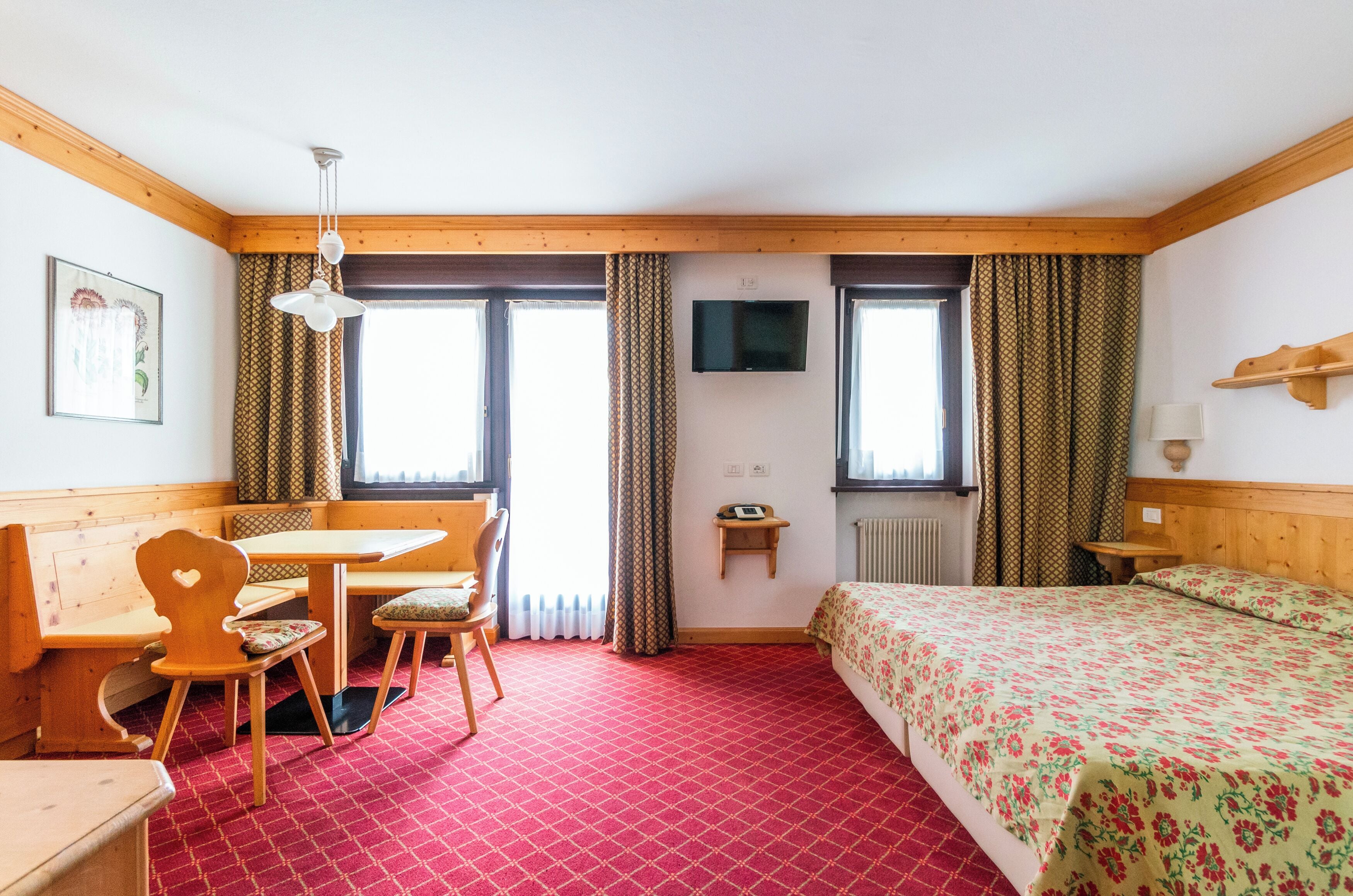 A hotel room with a wooden bench, a bed with floral bedding, a television, and a red patterned carpet.