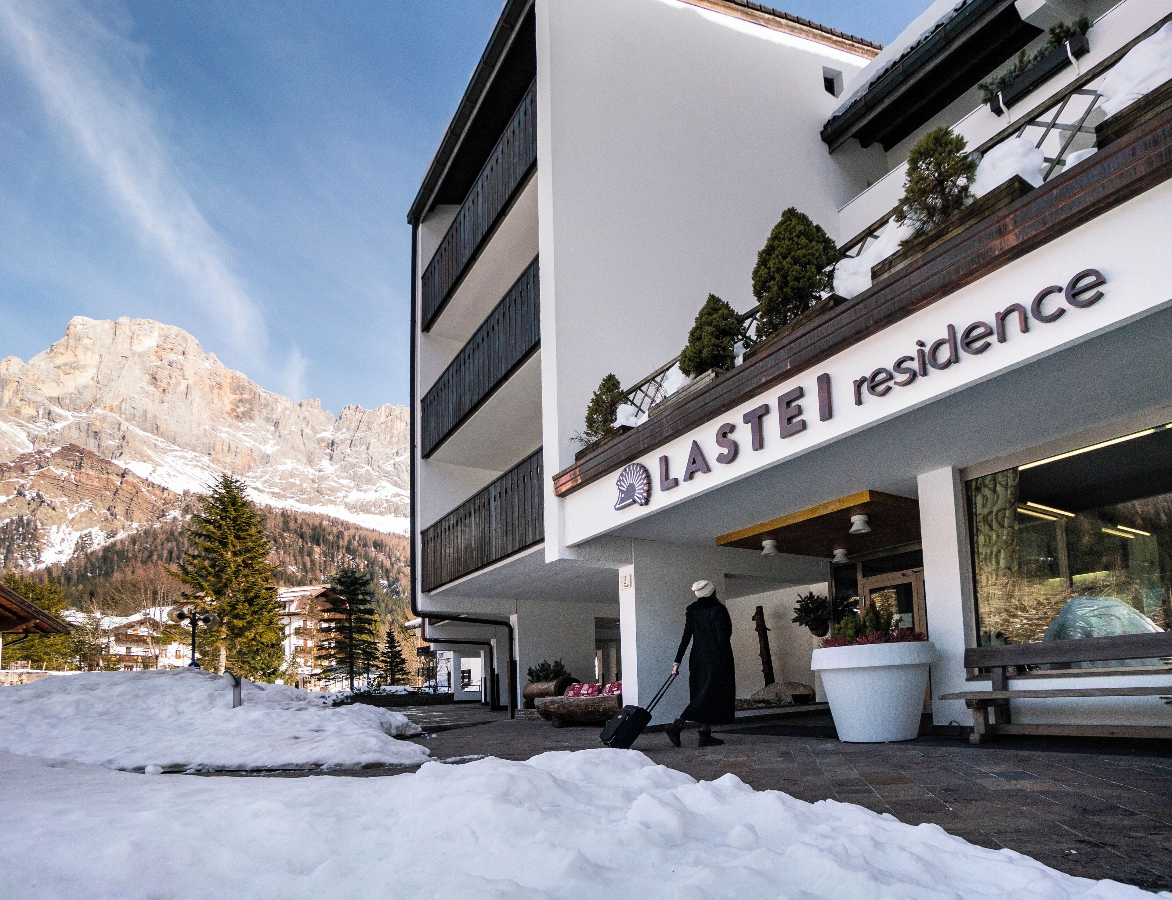 A hotel named Lastei Residence with a modern design, a person pushing a suitcase, and a snowy mountain backdrop.