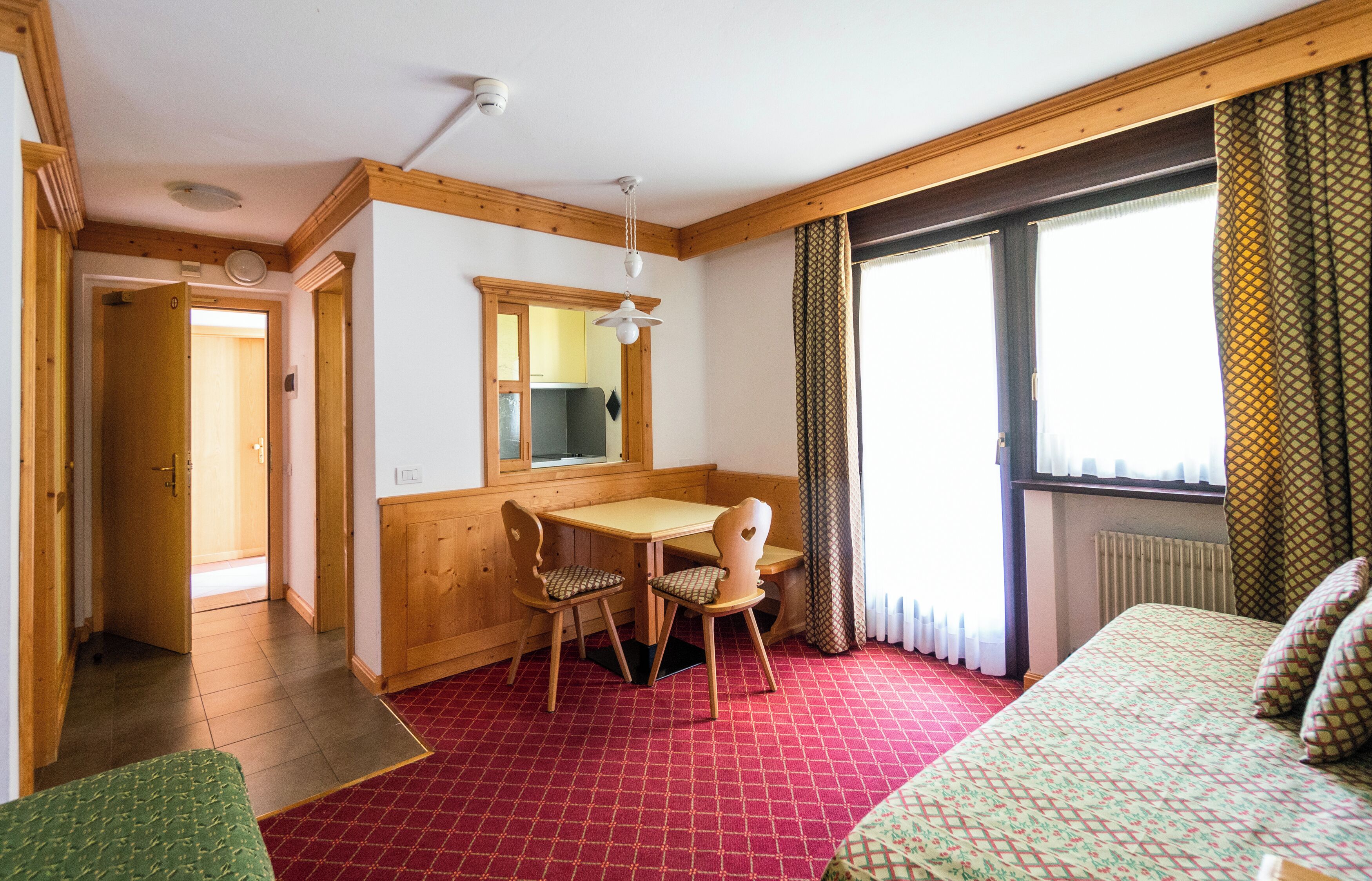 A room with a wooden table and chairs, a bed with patterned bedding, a window with curtains, and a radiator.