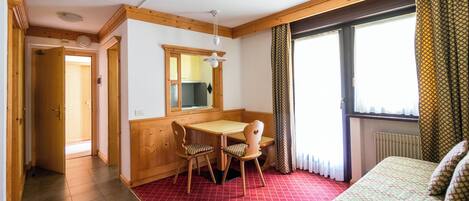 A room with a wooden table and chairs, a bed with patterned bedding, a window with curtains, and a radiator.
