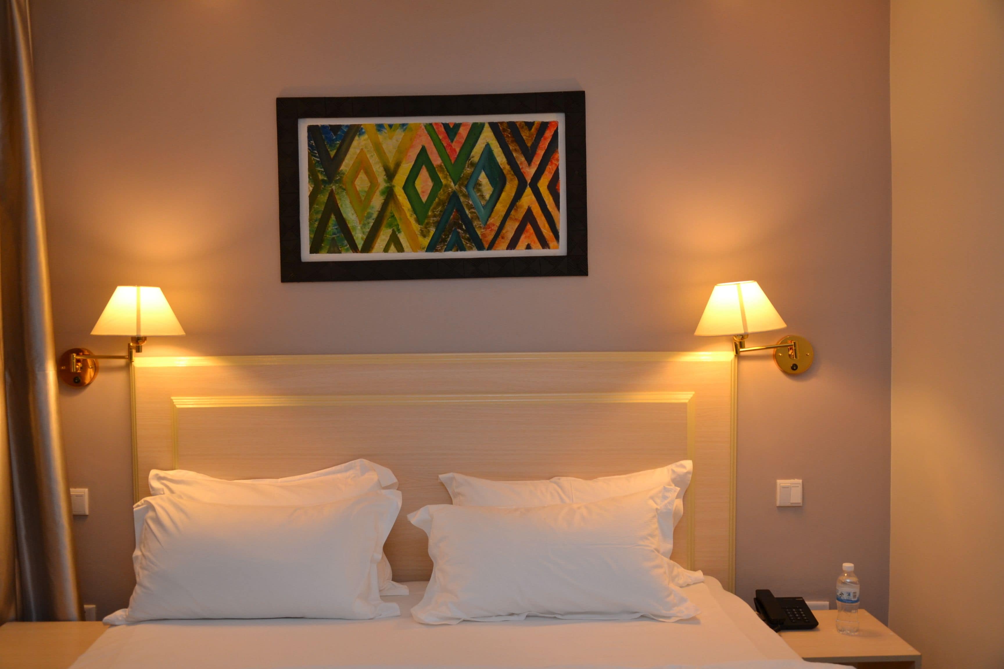 A hotel room with a bed, two bedside lamps, a framed artwork on the wall, and a water bottle on the nightstand.