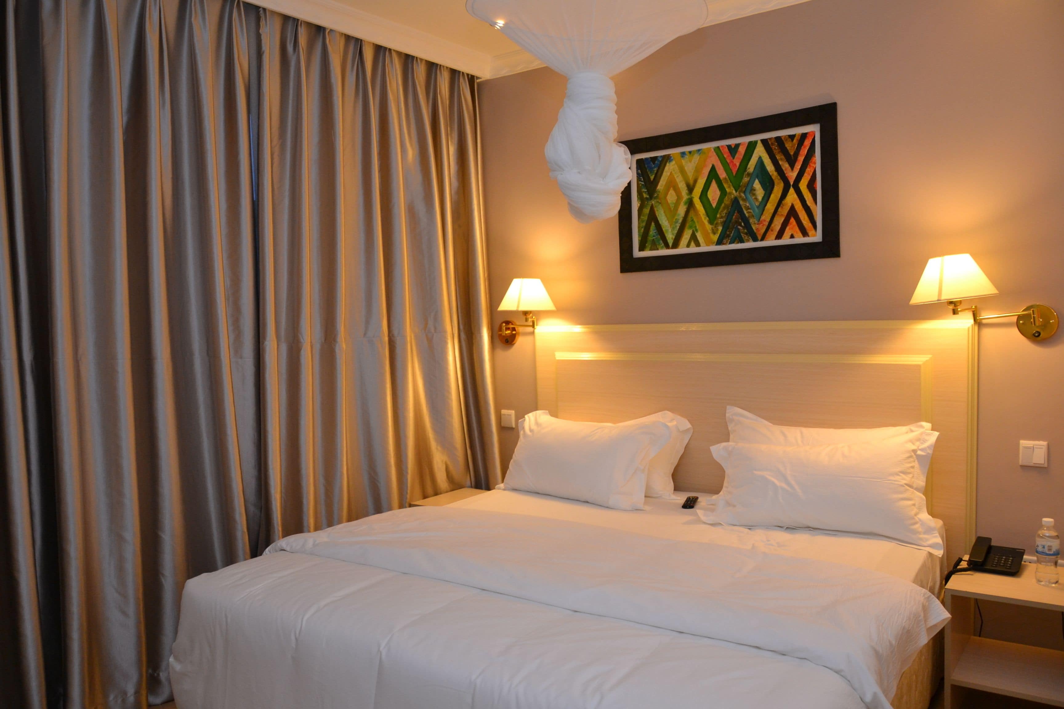 A hotel room with a large bed, two bedside lamps, a nightstand with a phone and a bottle, and a wall-mounted painting.