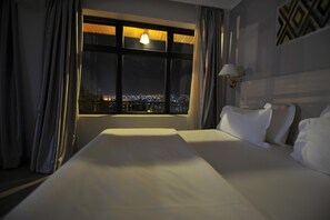 Une chambre d’hôtel avec un grand lit, une vue nocturne sur la ville par la fenêtre et une lampe sur la table de chevet.