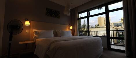 Ein Hotelzimmer mit einem großen Bett, Nachttischlampen, einem Standventilator und Blick auf die Stadt durch große Fenster.