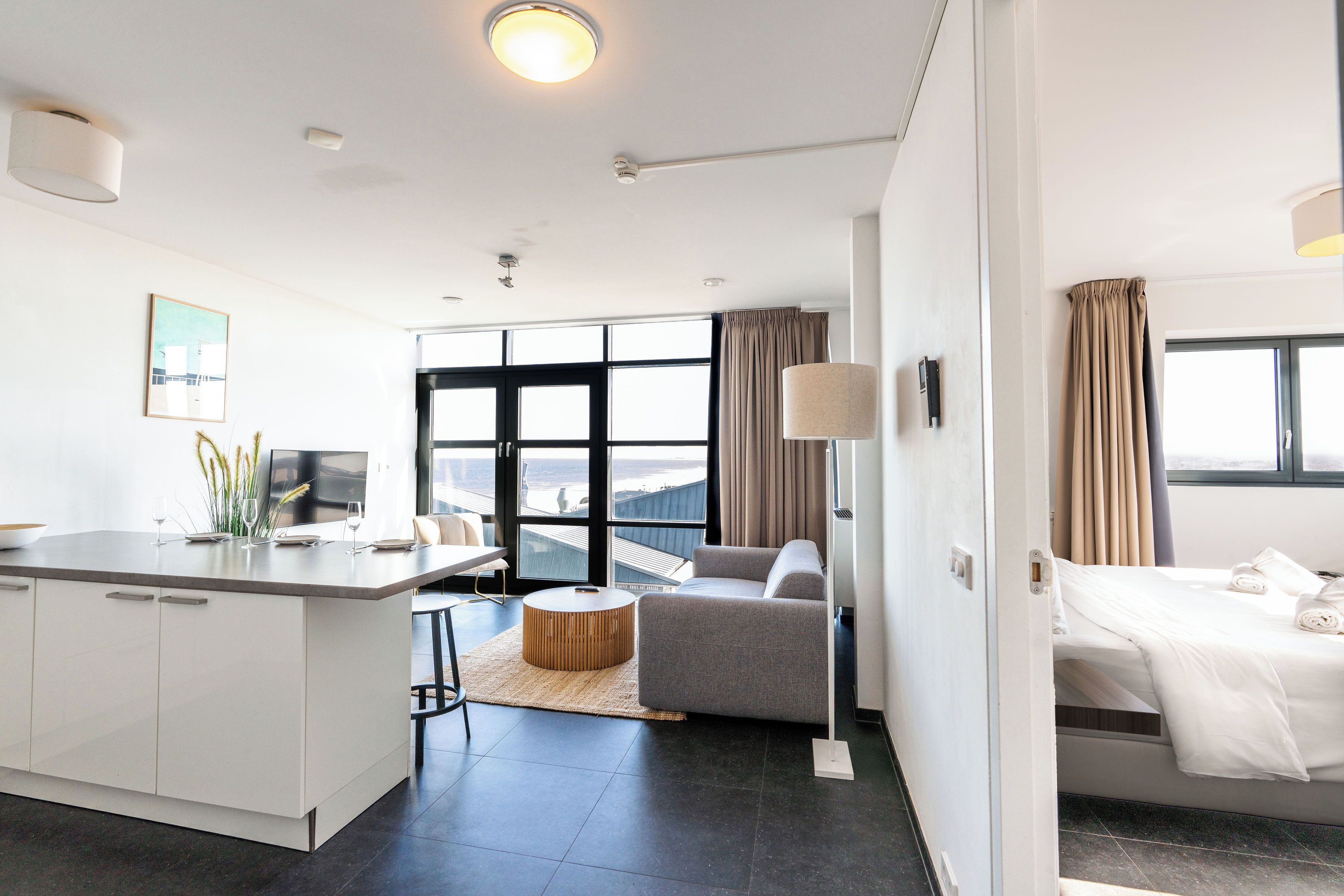 Ett modernt hotellrum med ett stort fönster, en soffa, en säng och ett litet köksutrymme.