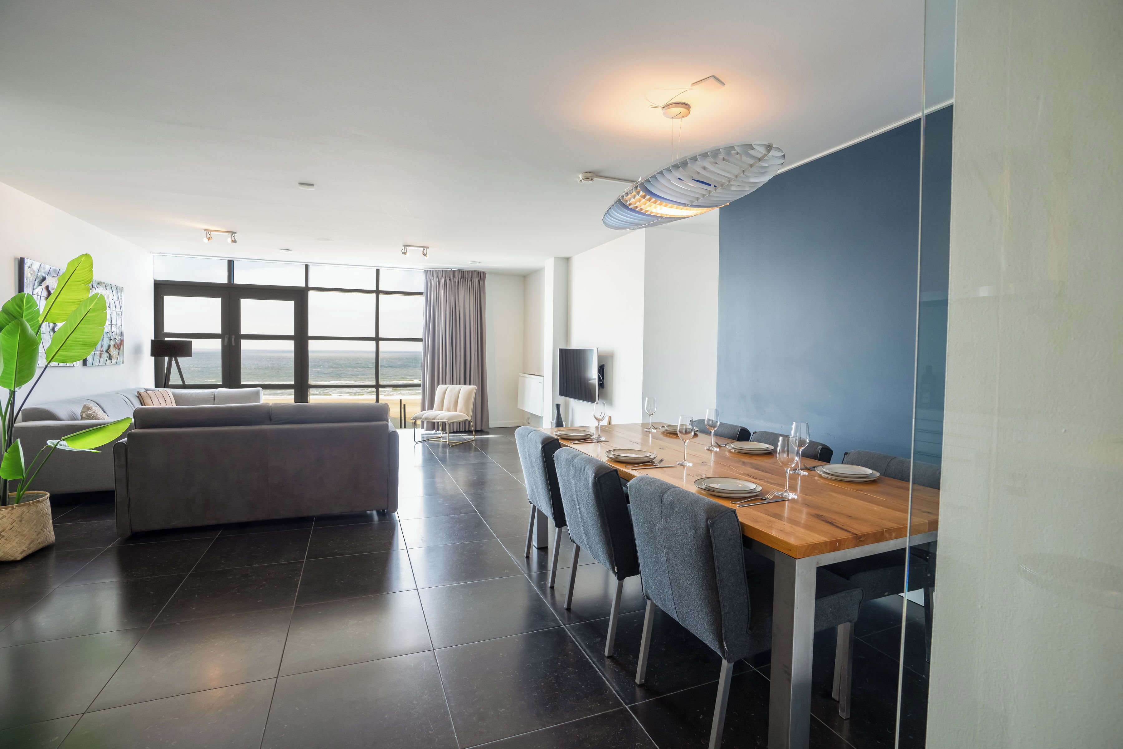 Een moderne eetruimte met een houten tafel voor vier personen, een grijze bank en een groot raam met uitzicht op de stad.
