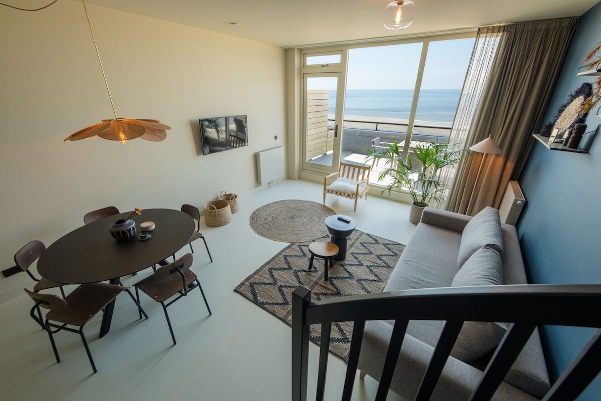 Una sala de estar moderna con comedor, una escalera y un amplio ventanal con vista al océano.
