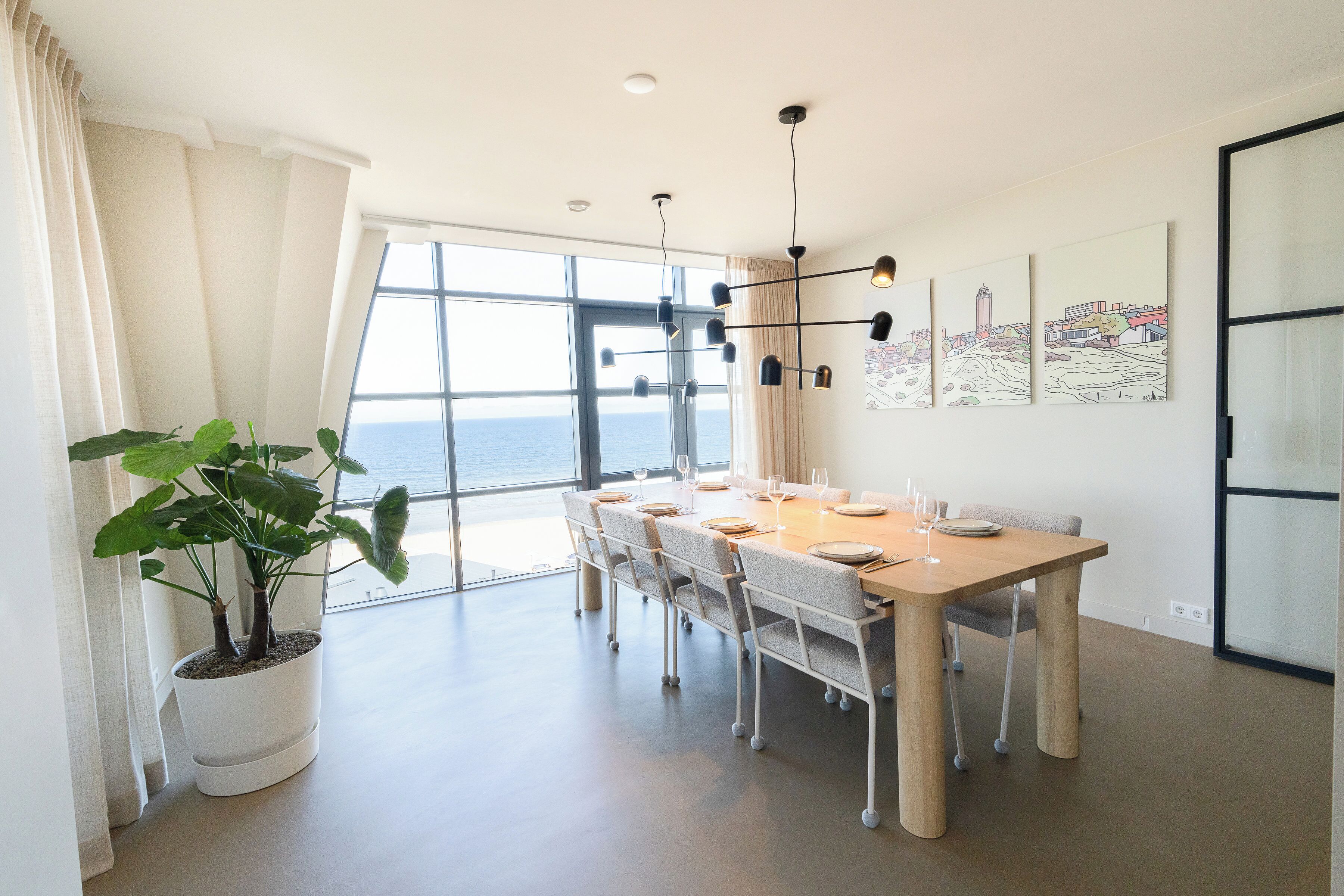 A modern dining area with a large table, chairs, a potted plant, and artwork on the walls.