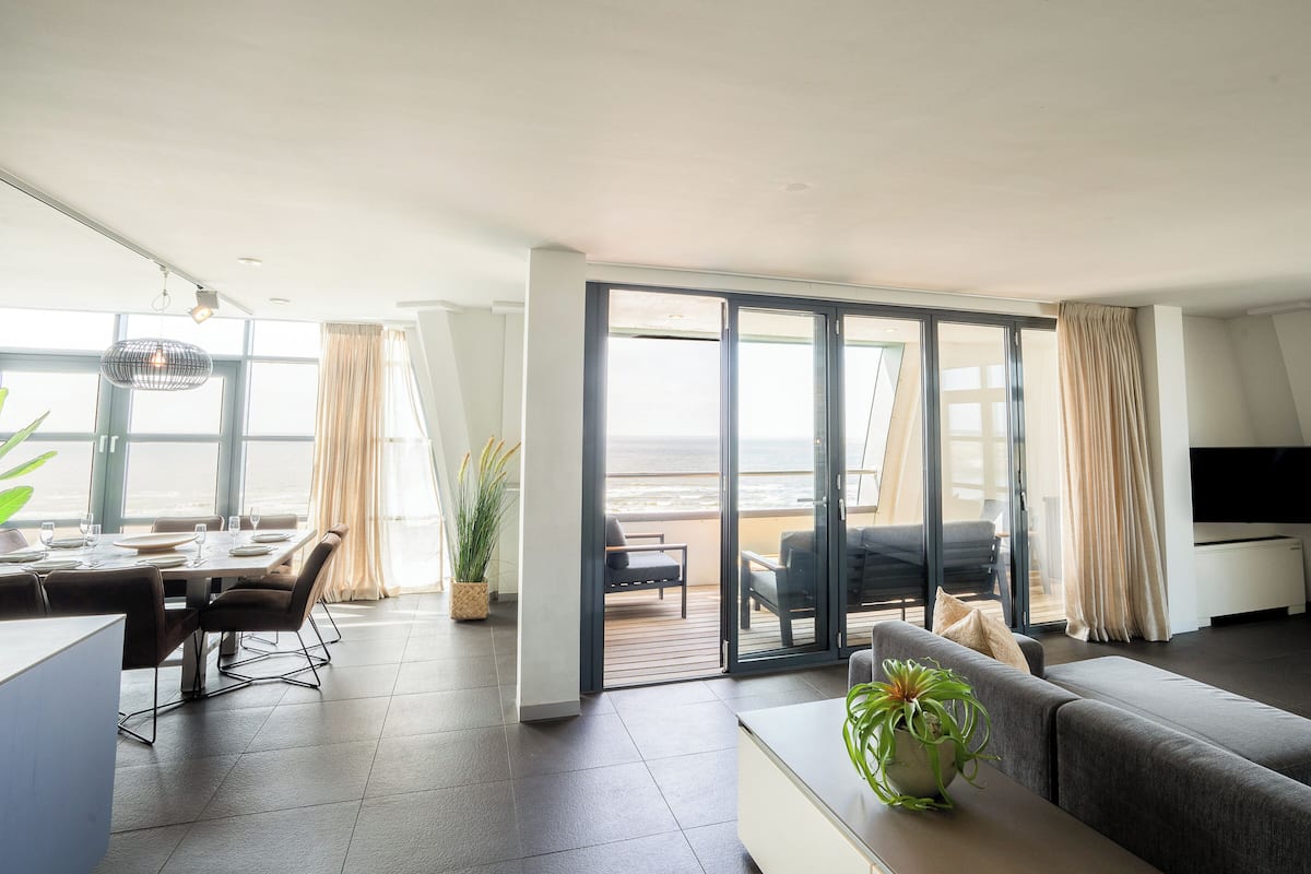 Una sala moderna con un sofá, un comedor con mesa y sillas, y un balcón con vista a la playa.