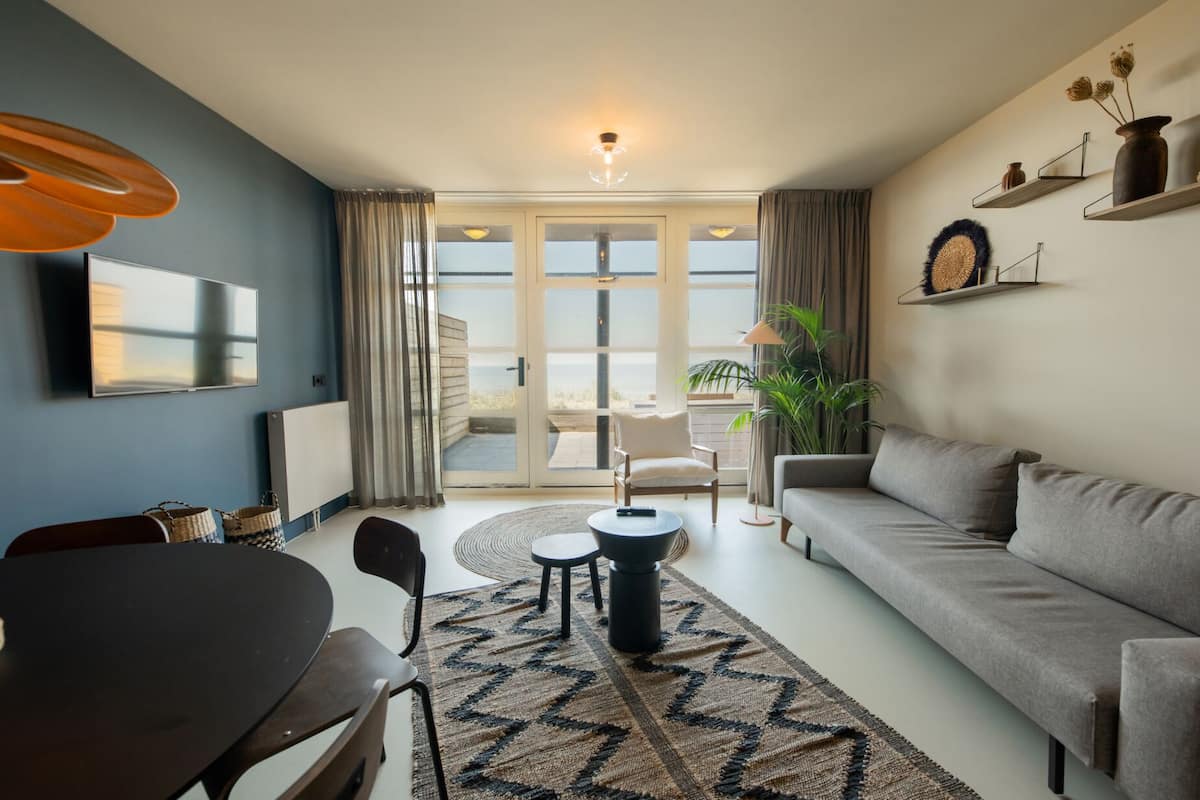 Una sala de estar moderna con un sofá, una mesa de comedor con sillas, un televisor y vistas al exterior.