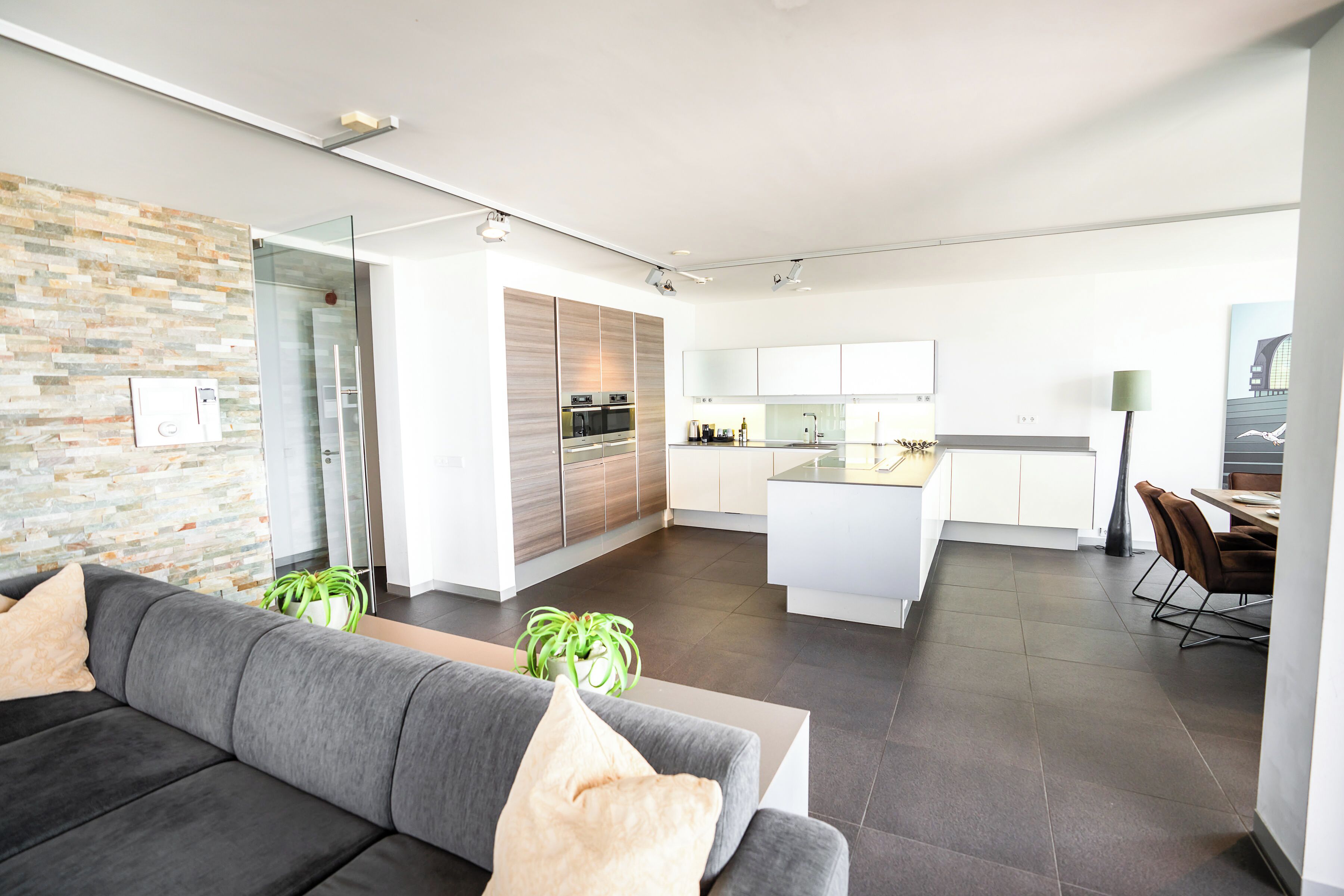 Una sala de estar moderna con un sofá gris, un puerta de vidrio que da a una cocina, y un comedor con una mesa y sillas.