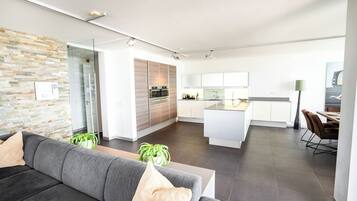 Ein modernes Wohnzimmer mit einem grauen Sofa, einer Glastür, die in eine Küche führt, und einem Essbereich mit Tisch und Stühlen.