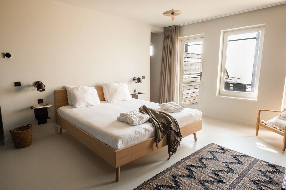 Un dormitorio con una cama grande, una mesita de noche, una lámpara, una ventana con cortinas y vistas al exterior.